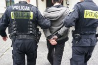 grafika- zdjęcie. dwóch policjantów prowadzi między sobą zatrzymanego mężczyznę, który ma kajdanki na rękach z tyłu. Policjanci trzymają  go za rękę w okolicy łokcia